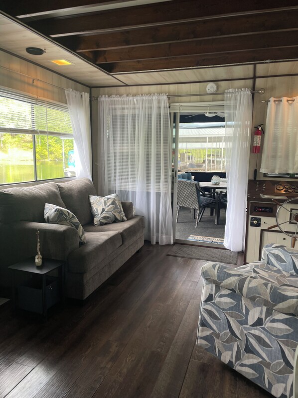 A cozy living area with a sofa, a patterned armchair, and a small table with a guitar. A window with curtains offers a view of the outdoors. - Houseboat Retreat on Norris Lake (Tazewell)