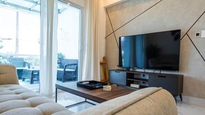 A modern living room with a large flat-screen TV, a sofa, a coffee table, and a sliding glass door leading to an outdoor area. - Tropico Smart by Suarez Properties | 2BR & Jacuzzi (Santo Domingo Este)