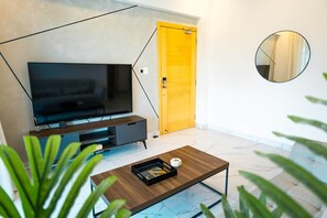 A modern living room with a flat-screen TV, a wooden coffee table, a yellow door, and a round mirror on the wall.
