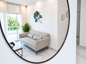 A modern living room with a sofa, a coffee table, and a view of greenery outside. - Tropico Smart by Suarez Properties 2BR & Jacuzzi (Santo Domingo Este)
