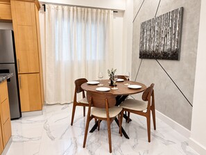 A dining area with a round table set for four, wooden chairs, a marble floor, a kitchen with wooden cabinets, and a large abstract painting on the wall.