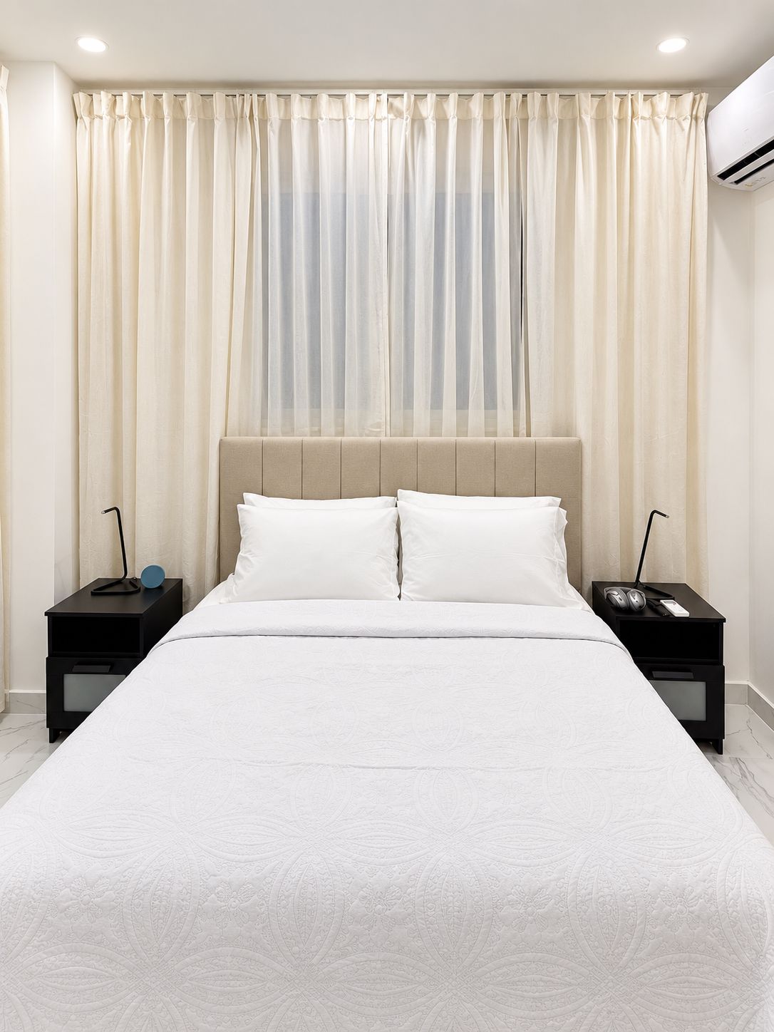 A neatly made bed with a headboard, two bedside tables with lamps, and sheer curtains.