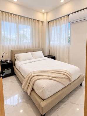 A modern bedroom with a large bed, a nightstand, an air conditioning unit, and sheer curtains.