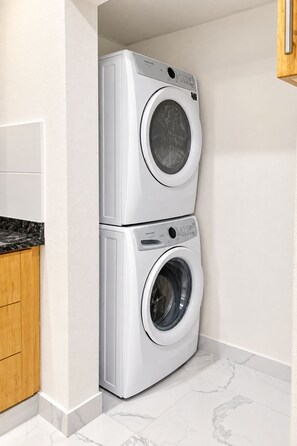 A compact laundry room with two stacked front-loading washing machines, wooden cabinets, and a marble-tiled floor.
