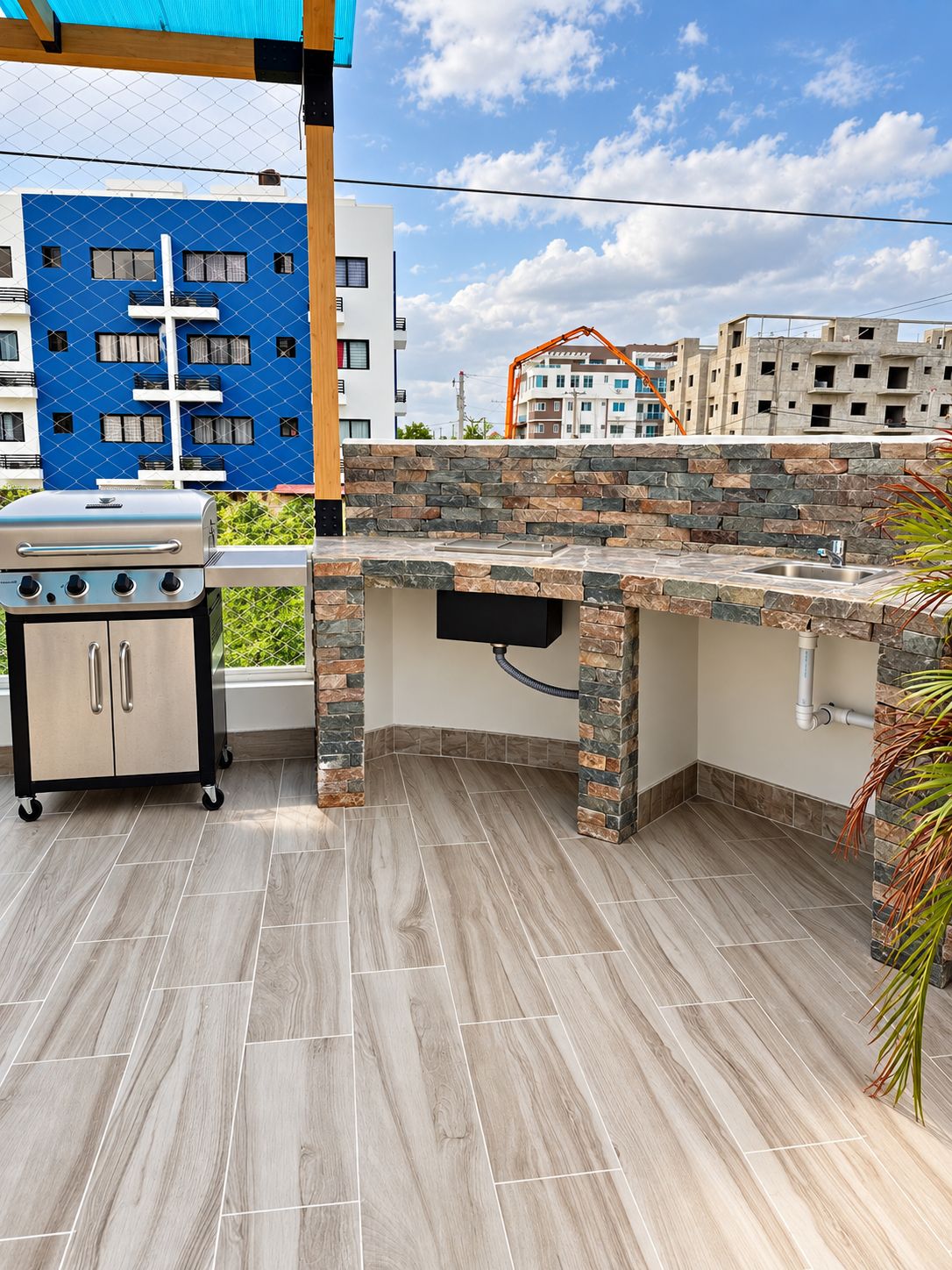 Un área de cocina exterior con una parrilla empotrada y un lavamanos, rodeada de detalles de piedra y vistas a edificios residenciales.