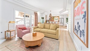 A spacious living room with a beige sofa, a pink armchair, and a round wooden coffee table. A large window provides natural light, and a kitchen is visible in the background. - Bright 3-bedroom townhouse, w/ocean views roof deck, AC, walk to everything! (San Diego)
