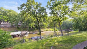 A riverside view with a fenced area, a small dock, and a building with a metal roof. - Lakefront Home on Grand Lake in Grove – Dock, 10 Beds, Game Room, Honey Creek (Grove)