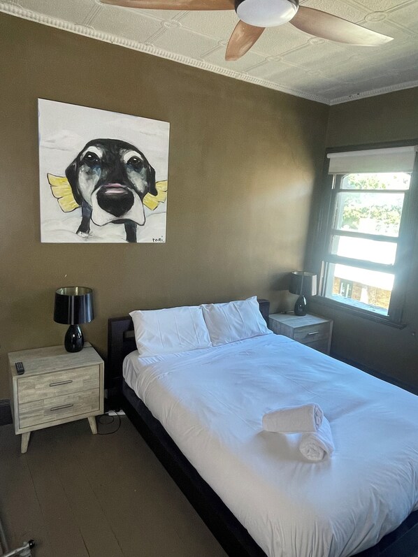A hotel room with a bed, a nightstand, a lamp, a painting of a dog, and a window with a view of greenery.
