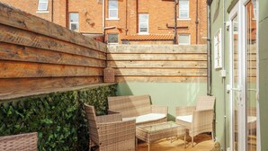 Une terrasse avec des meubles en osier, une plateforme en bois et, au fond, un bâtiment en briques.