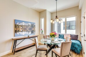 Uma sala de jantar com mesa de vidro, cadeiras e um lustre. Há um quadro na parede e uma janela com vista para o exterior.