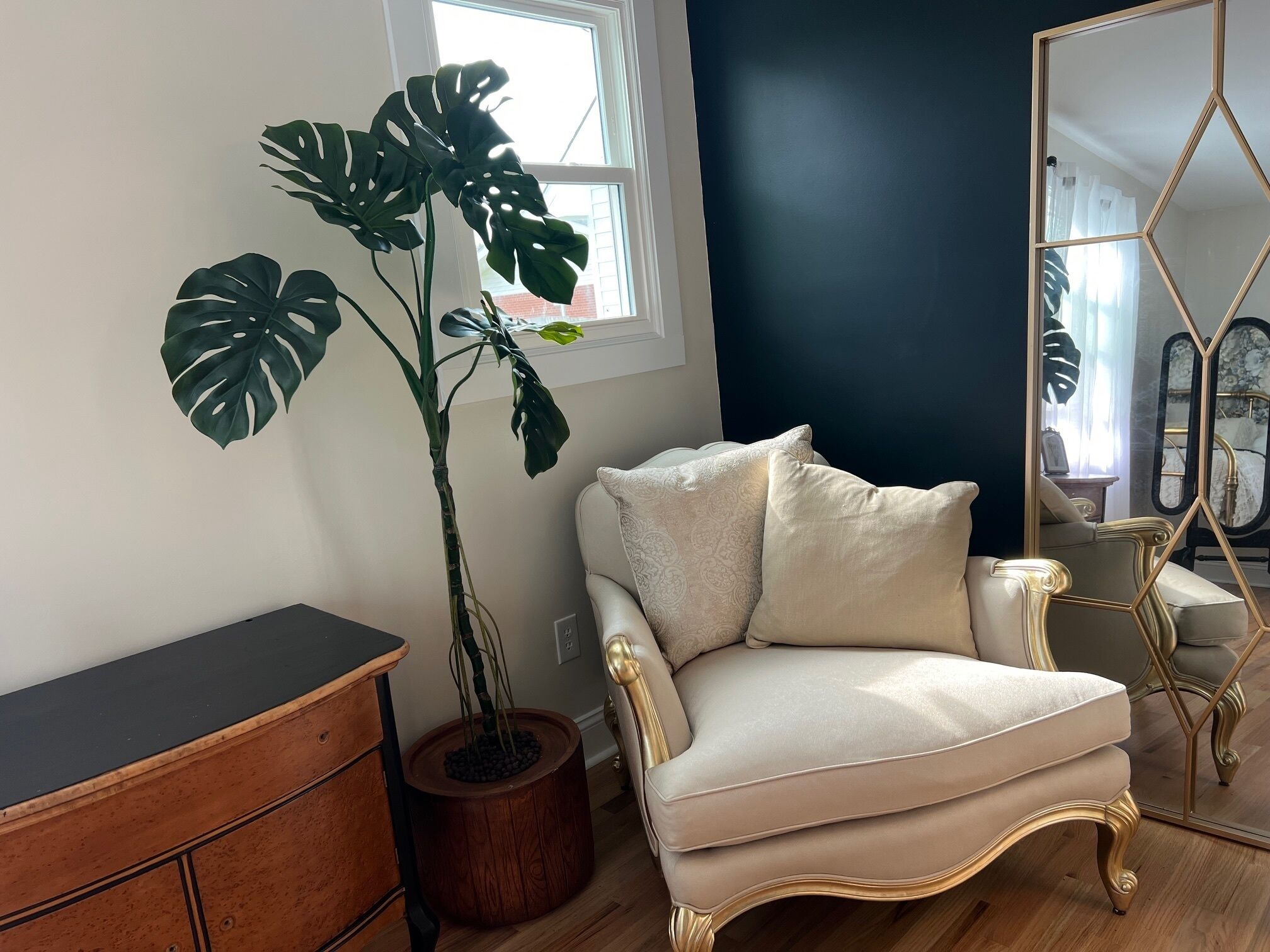 Een kamer met een grote potmonstera, een crèmekleurige fauteuil met gouden accenten, een houten dressoir en een spiegel met een geometrisch frame.