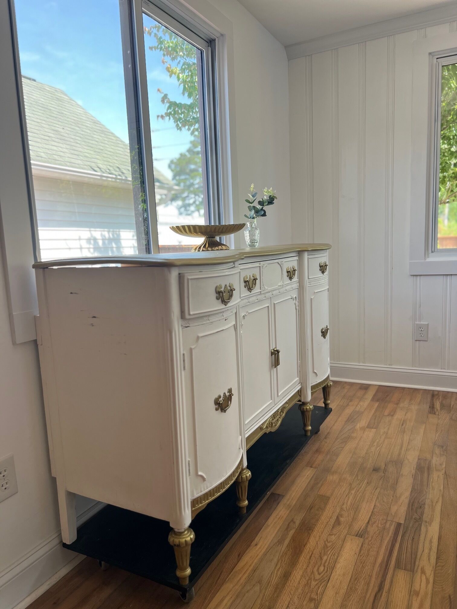Une commode blanche, richement décorée, avec des accents dorés et un socle noir, placée dans une pièce au plancher en bois et aux murs blancs.