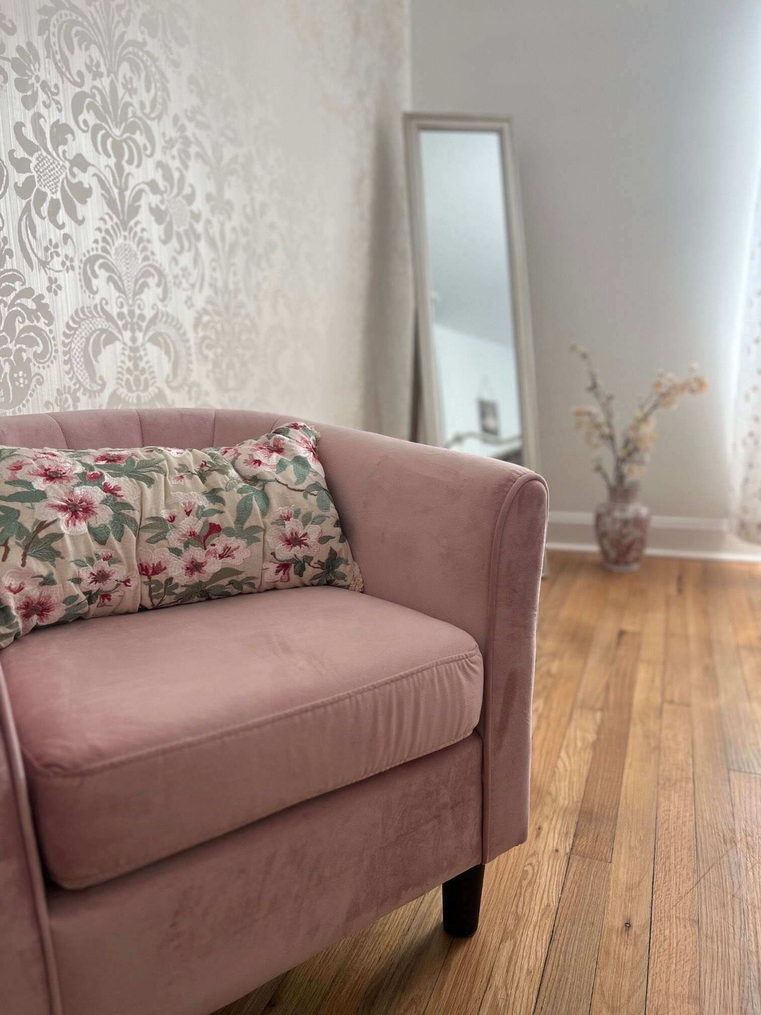 Un canapé rose agrémenté d’un coussin à motifs floraux, dans une pièce dont le papier peint est orné de motifs, avec un miroir et un vase de fleurs.