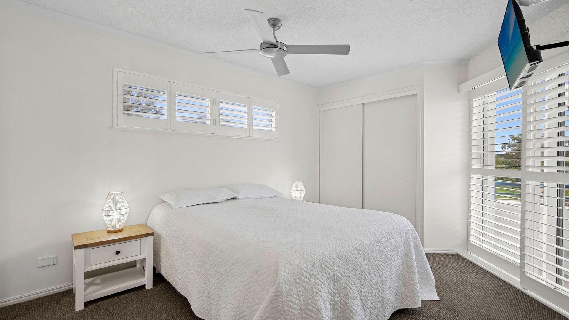 A bedroom with a bed, nightstand, lamp, ceiling fan, and window blinds.