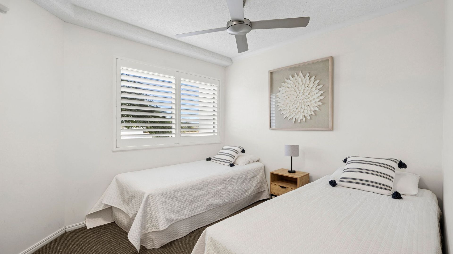 A room with two single beds, a ceiling fan, a window with blinds, a bedside table with a lamp, and a wall art piece.