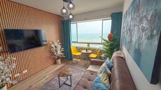 A living room with a TV, wooden panel wall, sofa, chairs, and a view of the sea.