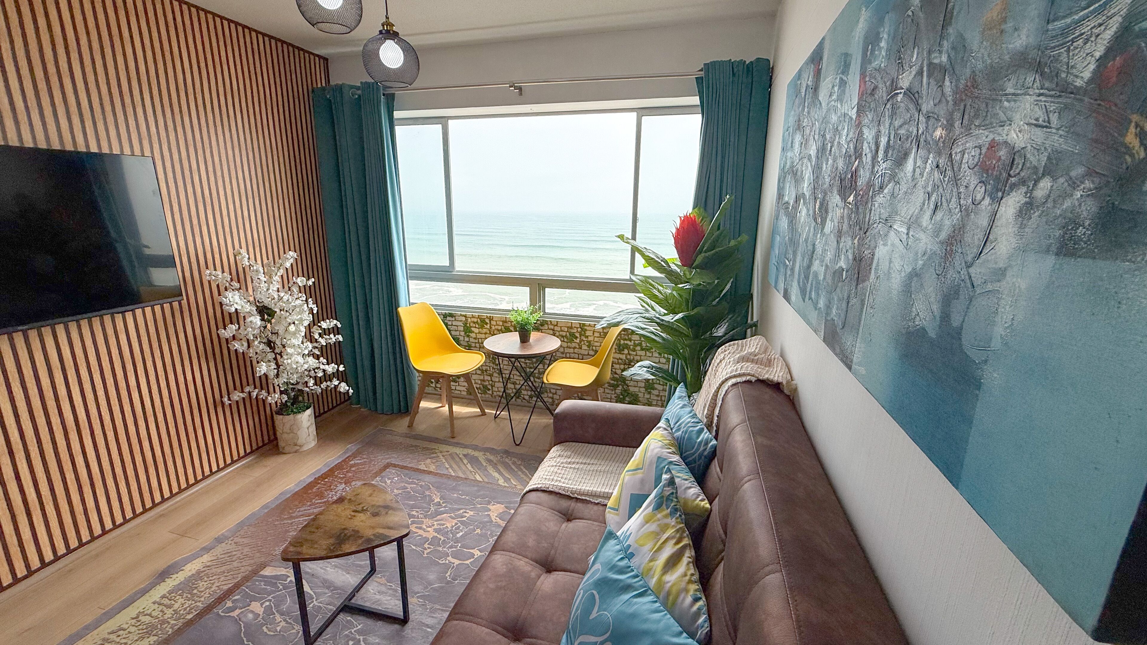 A modern living room with a large window, a flat-screen TV, a brown sofa, yellow chairs, and a potted plant.