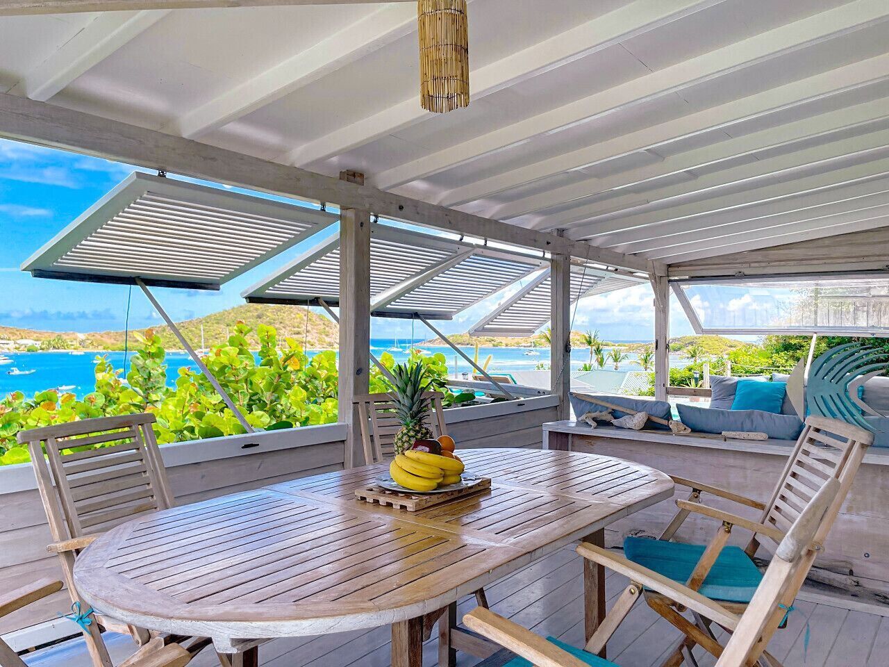 Un'area pranzo all'aperto coperta con tavolo e sedie in legno, una ciotola di frutta e vista sull'oceano e sul verde.