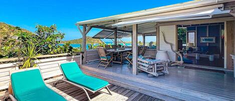 A wooden deck with two green lounge chairs, a covered outdoor dining area, and a view of the ocean.