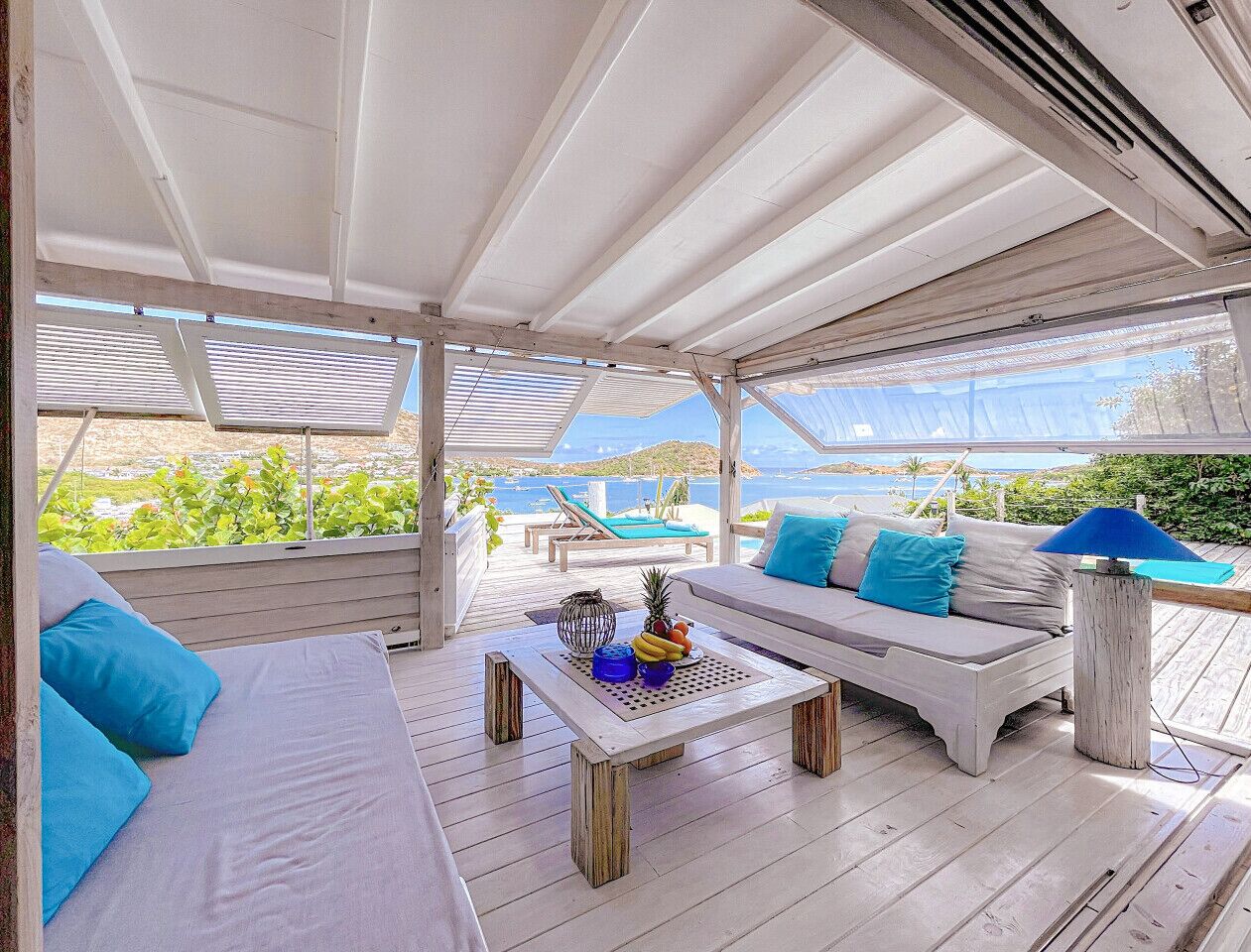 Un espacio de estar al aire libre techado con un sofá y una mesa, una piscina al fondo y vistas al mar.