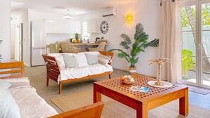 A modern living room with a wooden sofa, a coffee table, and a dining table. There is a kitchen in the background with white cabinets and a refrigerator. A large window with curtains is visible, and a potted plant is on the windowsill. - Villa Côté Sable, Accès plage, Vue mer, Elégance (Orient Bay)