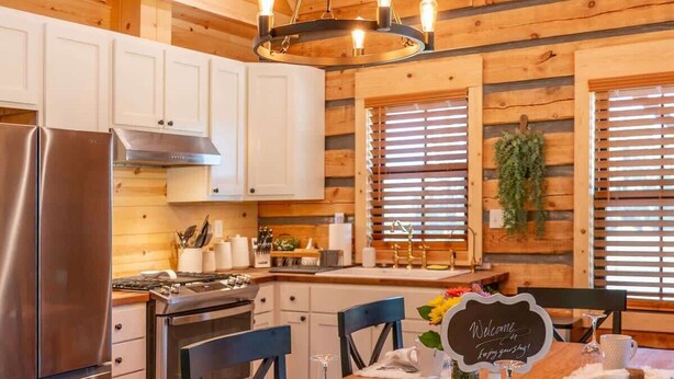 Ein Holzblockhaus-Interieur mit einem Esstisch, gedeckt für vier Personen, einem Edelstahl-Kühlschrank und einer modernen Küche mit weißen Schränken und einer hängenden Pflanze.