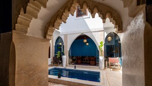 Una piscina interior con suelo de baldosas, rodeada de arcos y columnas, y equipada con sillas y mesas.