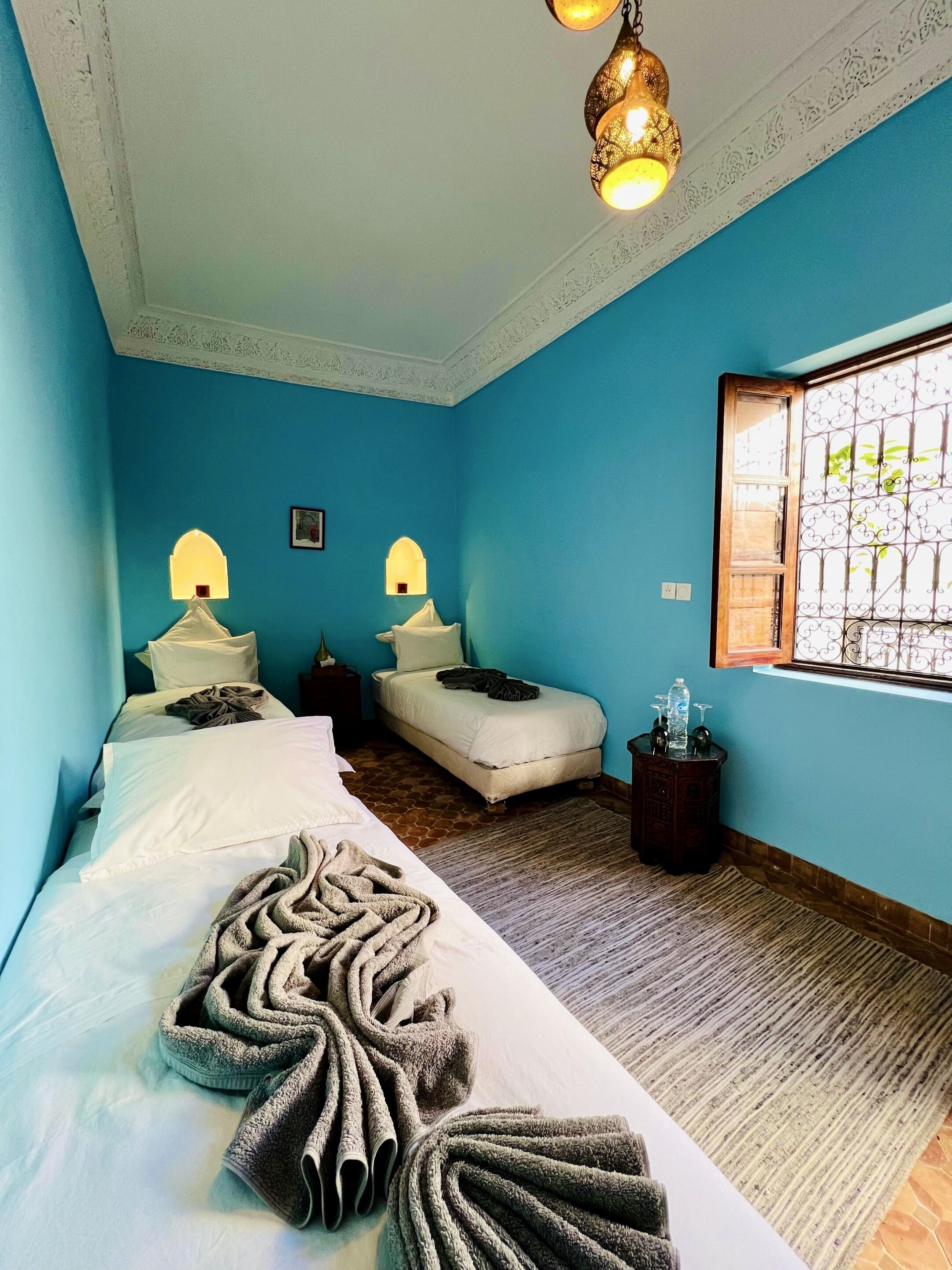 A bedroom with two beds, a patterned rug, a window with decorative iron bars, and a hanging light fixture.