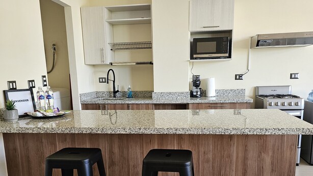 A kitchen with a granite countertop, two black barstools, a microwave, a gas stove, and a sink.