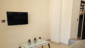 A modern living room with a flat-screen TV mounted on the wall, a glass coffee table with three potted plants, and a beige sofa.