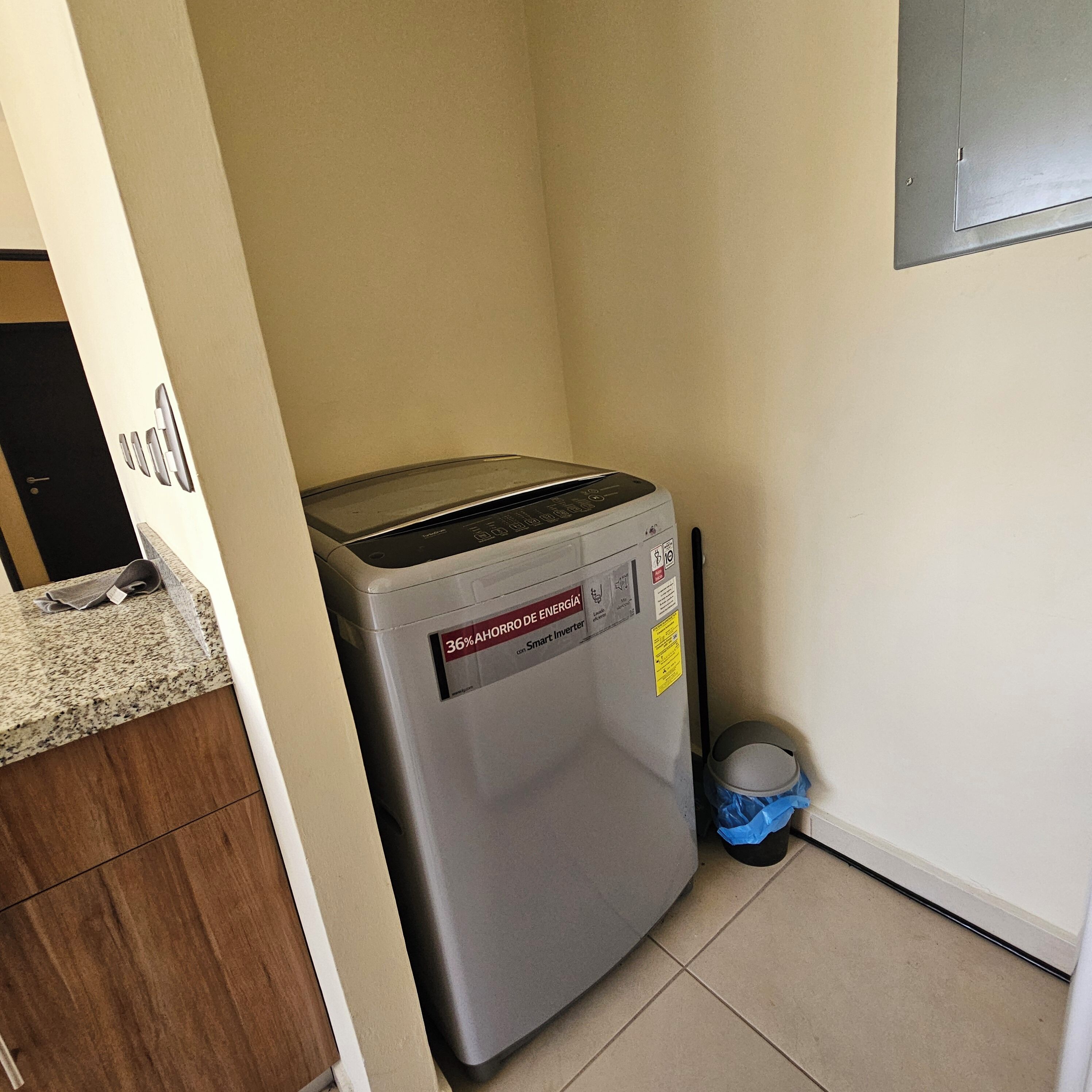 Una lavadora compacta de color gris, de carga frontal y con pantalla digital, ubicada en una pequeña área de cocina con suelo de baldosas y un mueble de madera.