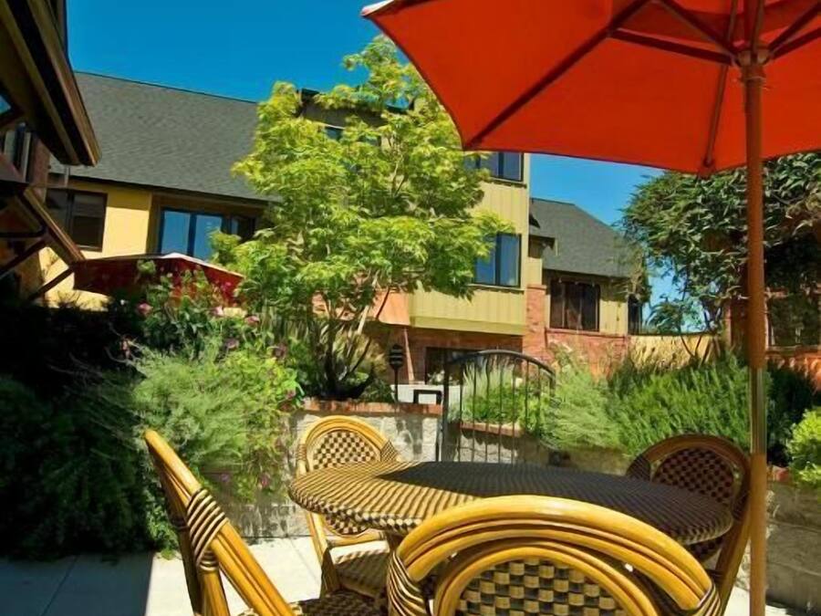 An outdoor seating area with wicker chairs and a table under a red umbrella, surrounded by greenery and buildings.