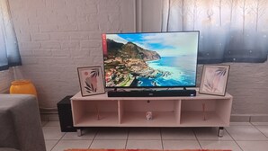 A living room with a flat-screen TV on a white entertainment unit, a patterned rug, and a small table with a vase of flowers. - Close Curtains Maboneng (Johannesburg)