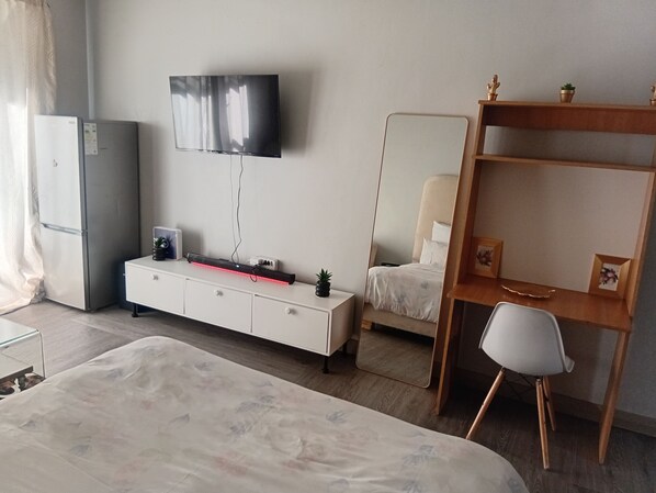 A hotel room with a bed, a desk, a chair, a TV, a wardrobe, and a small plant.