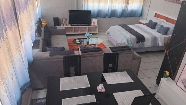 A modern hotel room with a sofa, a bed, a dining table with chairs, a TV, and a small coffee table. - Close Curtains Maboneng (Johannesburg)