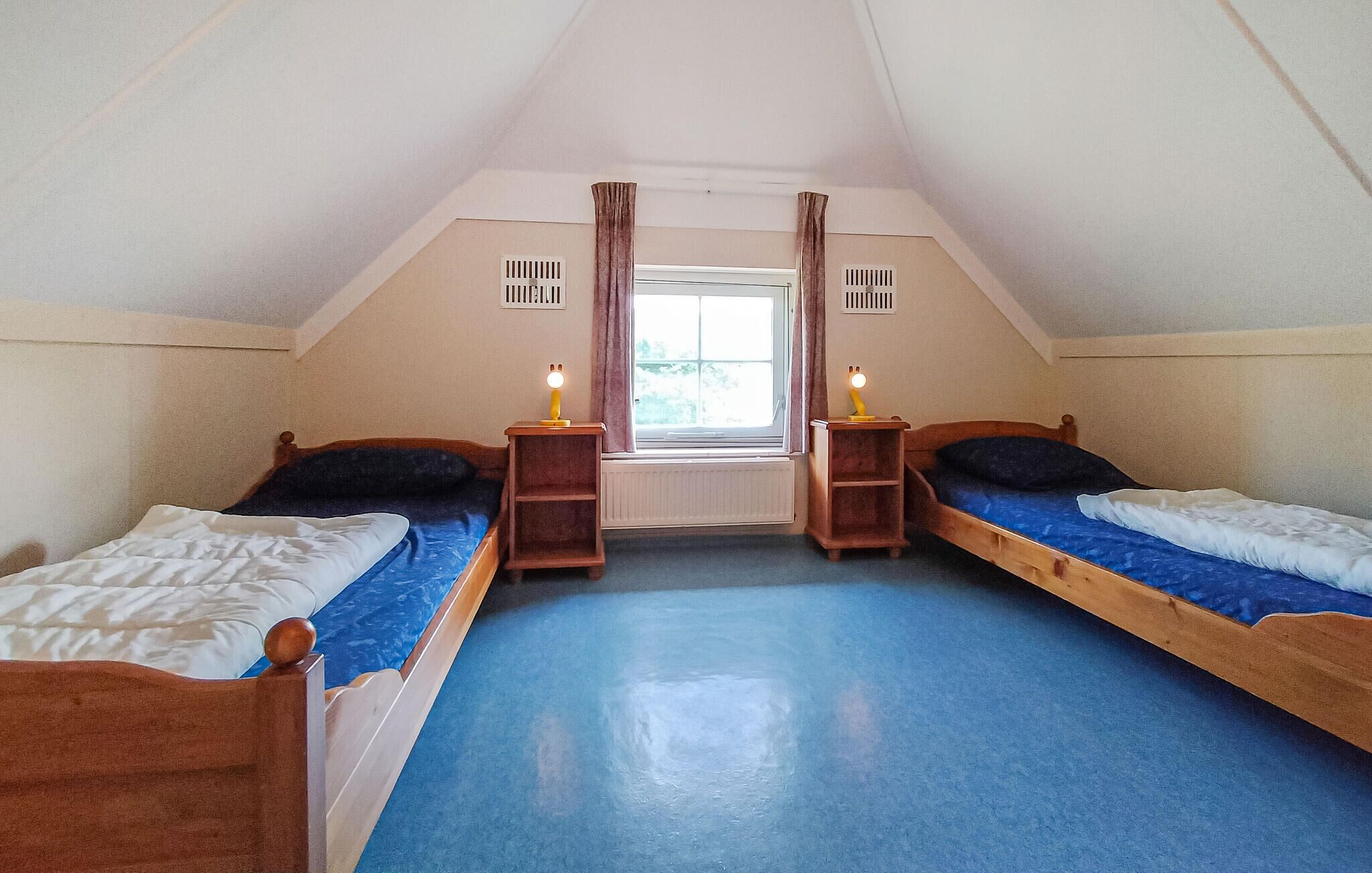 Une petite chambre mansardée triangulaire, avec deux lits simples, une fenêtre avec des rideaux et un radiateur.