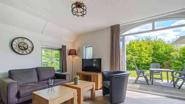 Un salon moderne avec un canapé, une table basse, un téléviseur et une vue sur un jardin verdoyant grâce à de grandes fenêtres.