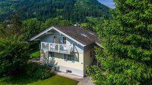 A two-story house with a balcony, surrounded by lush greenery and mountains in the background.