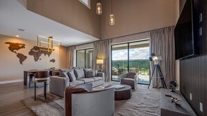 A spacious living room with a large sofa, a coffee table, and a TV. There is a world map on the wall and a large window with curtains. - Modern 3BR Disney Retreat | Jacuzzi & BBQ Patio (Kissimmee)
