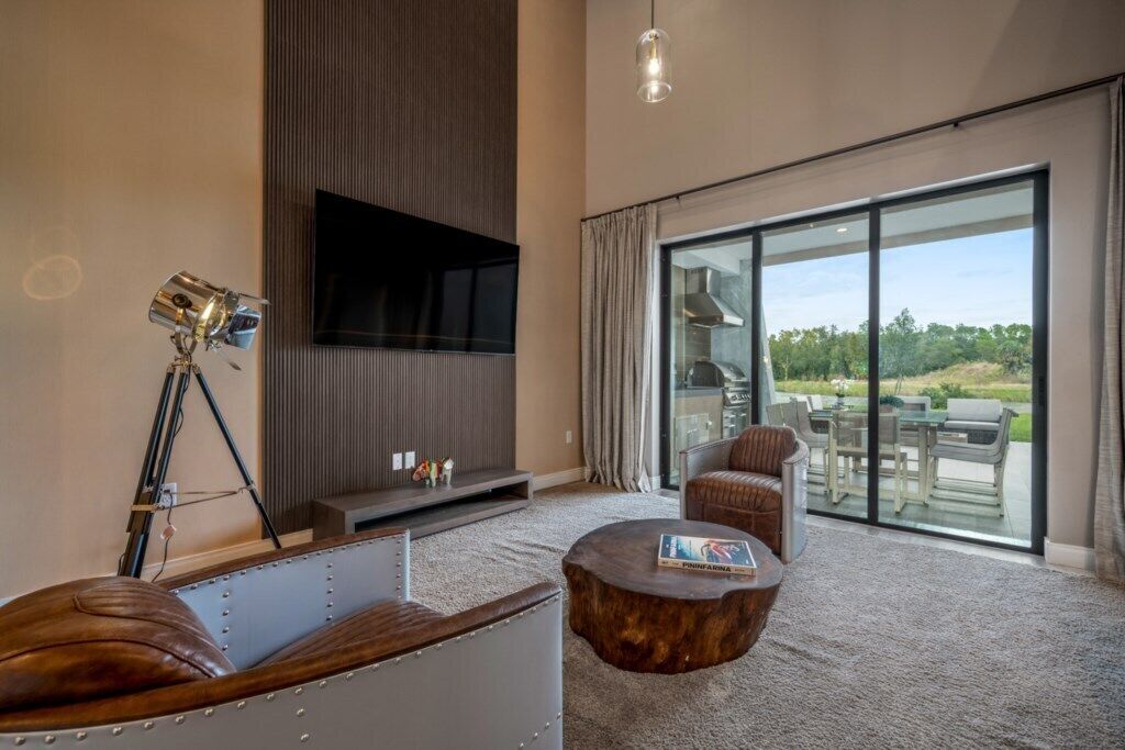 Een moderne woonkamer met een groot flatscreen televisietoestel, een op een statief gemonteerde camera, een leren bank, een ronde salontafel en een grote schuifdeur die leidt naar een buitenpatio.