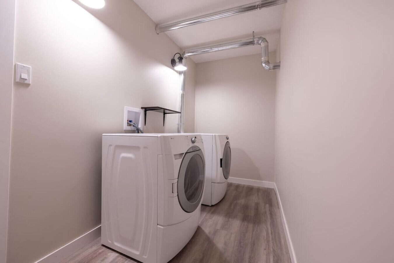 A laundry room with two front-loading washing machines, a light fixture, and a vent pipe on the ceiling.