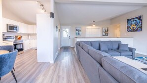 A modern living space with a grey sectional sofa, a kitchen with white cabinets, and a dining area with a blue chair.