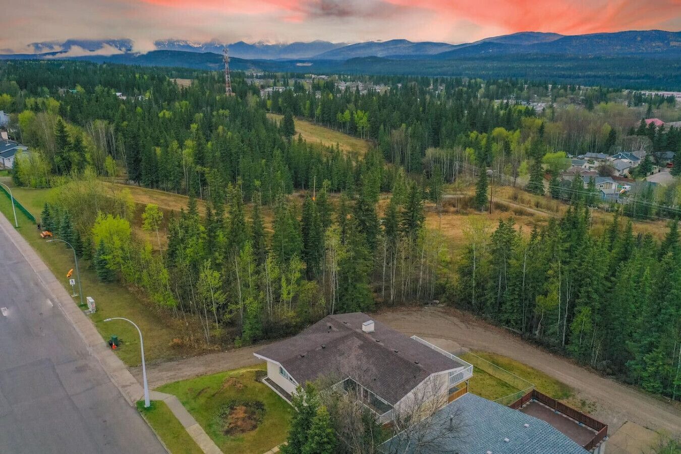 A residential area with a house surrounded by dense forest, a road, and a distant mountain range.