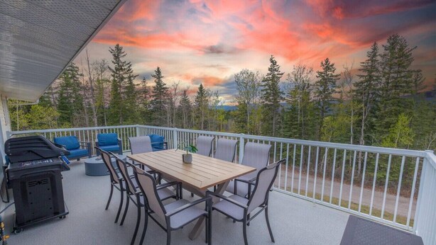 A covered outdoor patio with a wooden table and chairs, a grill, and a view of trees and a sunset.