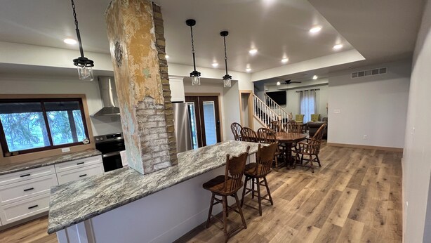 A spacious kitchen with a large island, wooden dining table, and chairs. There is a stone accent wall and a view of the outdoors through a window.