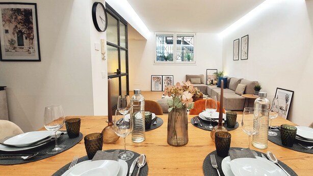 Ein Essbereich mit einem Holztisch, gedeckt für vier Personen, umgeben von Stühlen. An den Wänden befinden sich gerahmte Bilder, im Hintergrund ein Sofa und ein Fenster mit Blick.