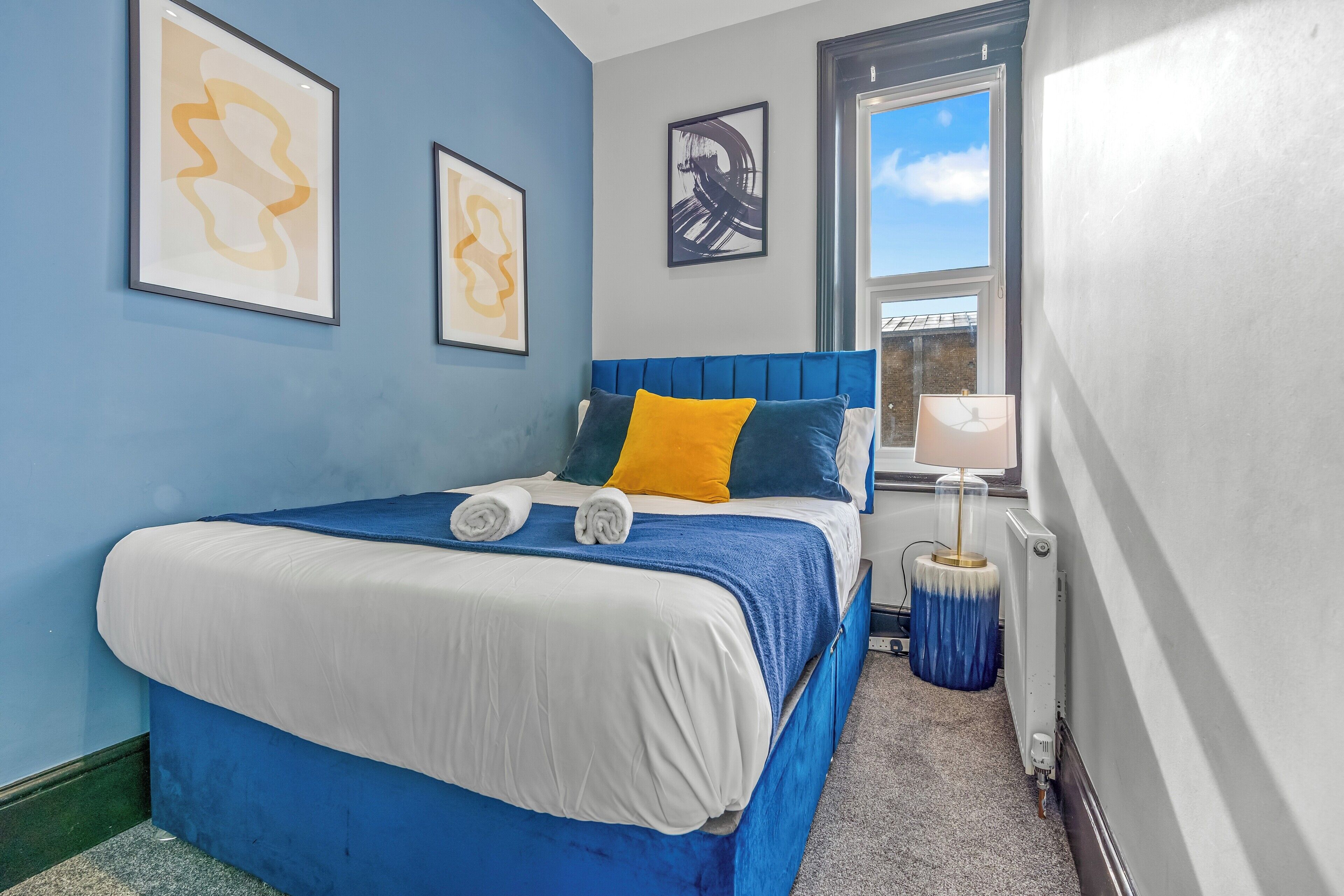 Un dormitorio con cabecera azul, cama con ropa de cama blanca y azul, almohada amarilla y dos cuadros en la pared.