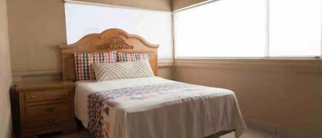 A bedroom with a wooden bed, a nightstand, a window with blinds, and a tiled floor.