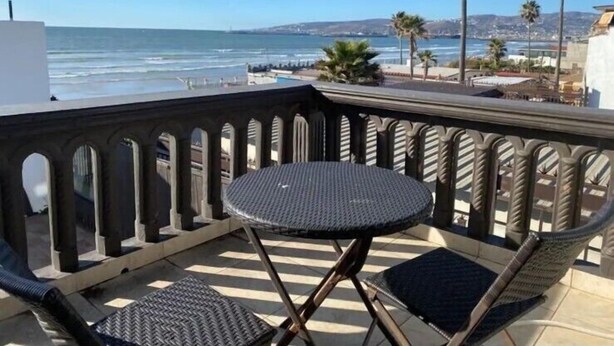 Un balcon avec une table ronde et deux chaises, donnant sur une plage bordée de palmiers et sous un ciel dégagé.