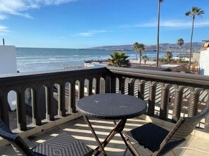 Un balcone con un tavolo rotondo e due sedie, con vista su una spiaggia con palme e cielo sereno.